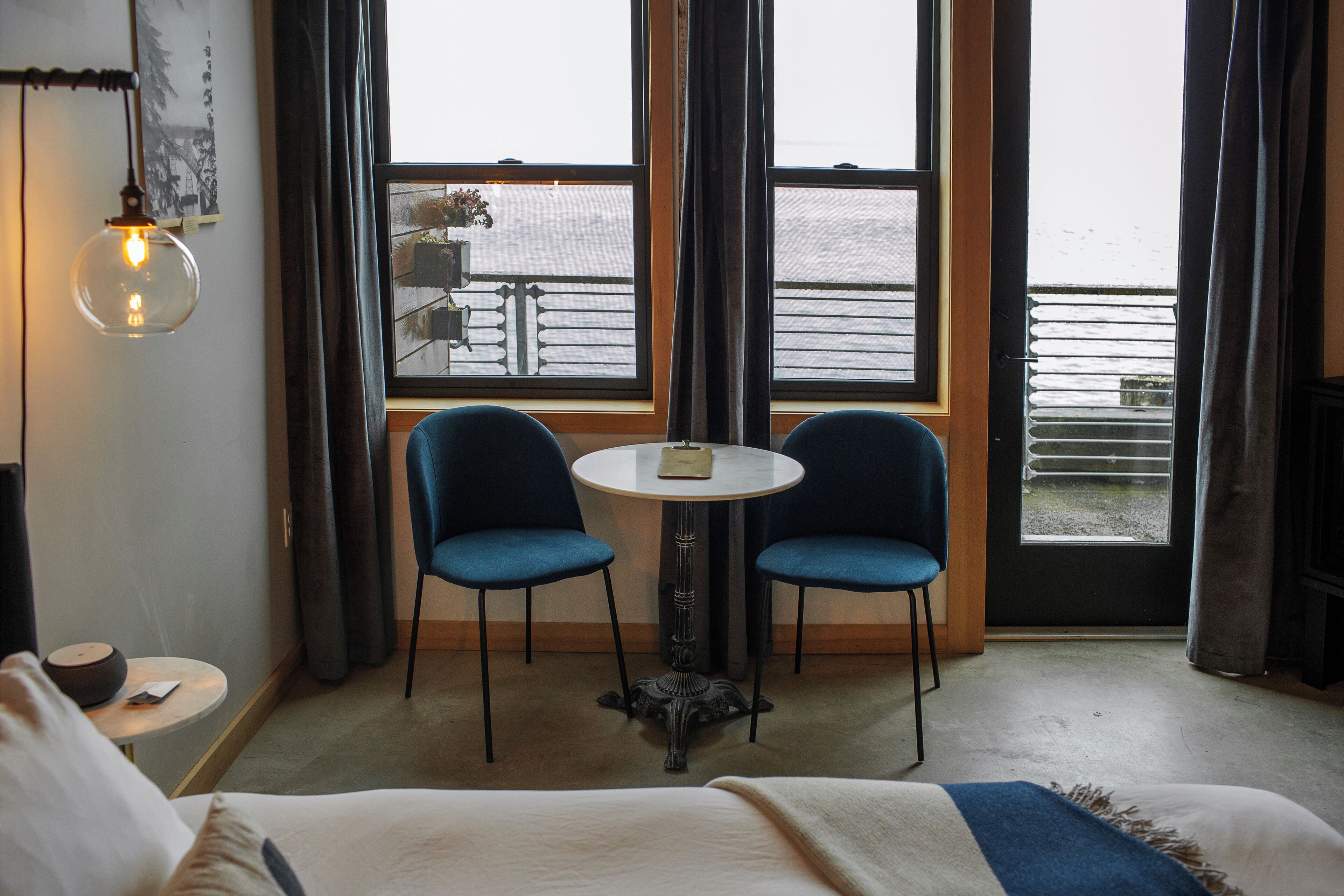 Ett hotellrum med en säng, ett litet runt bord med två stolar, en väggmonterad lampa och ett fönster med utsikt över ett snötäckt landskap.