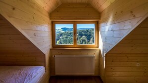 A wooden cabin room with a sloped ceiling, a double window with a forest view, and a white radiator.
