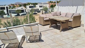 Une terrasse dotée de meubles en osier, d’un sol carrelé et offrant une vue sur des bâtiments et des montagnes en arrière-plan.