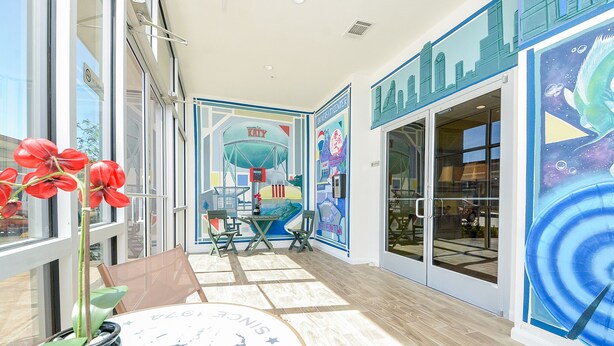 Un espacio interior moderno con puertas de vidrio, una mesa con un objeto decorativo y un mural que muestra un paisaje urbano y un pájaro.