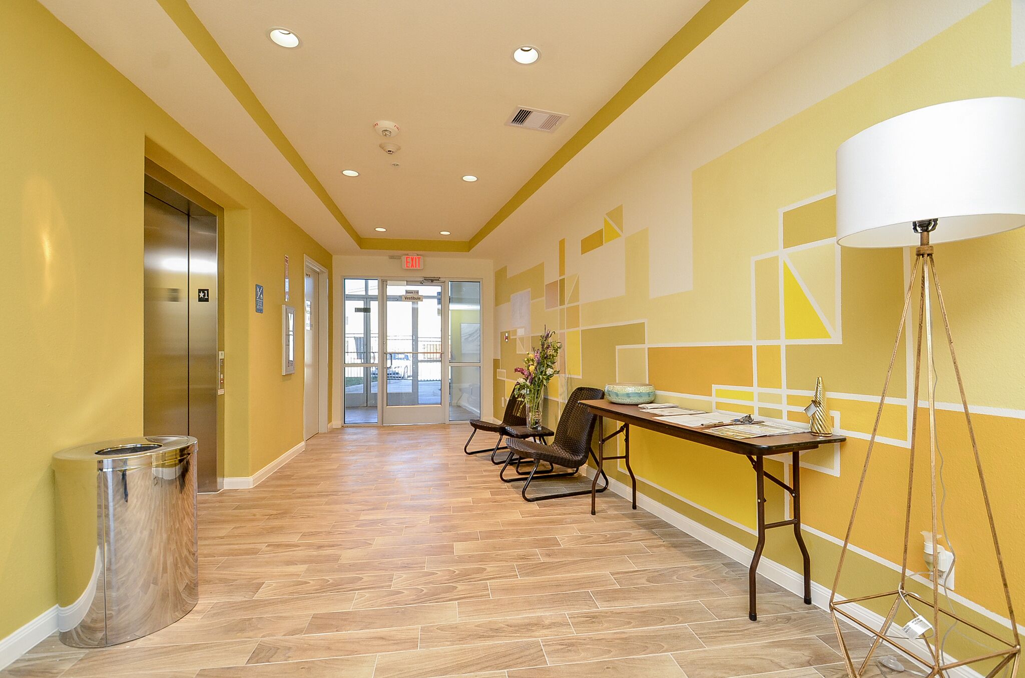 A hallway with an elevator, a trash can, a reception desk, and a lamp.