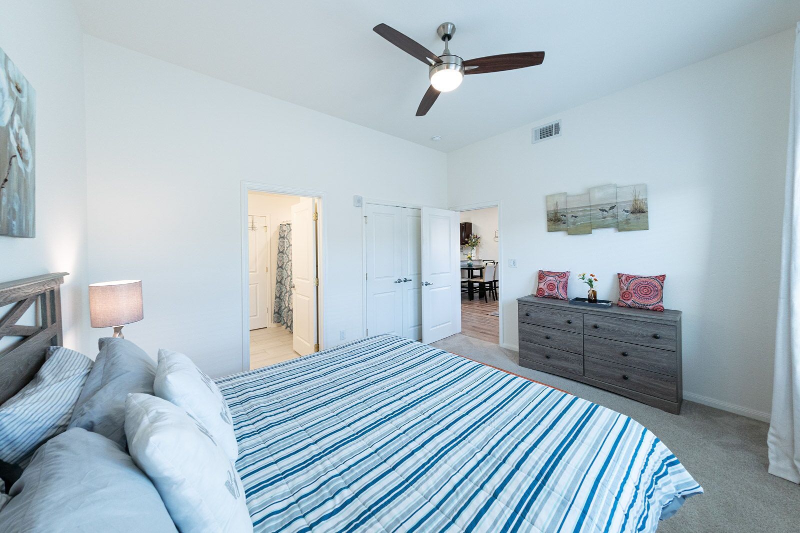 A bedroom with a bed, a dresser, a ceiling fan, and a door leading to a dining area.