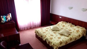 A hotel room with a bed, a desk with a television, a nightstand, and a window with curtains. - Oxa Hotel (Novi Pazar)
