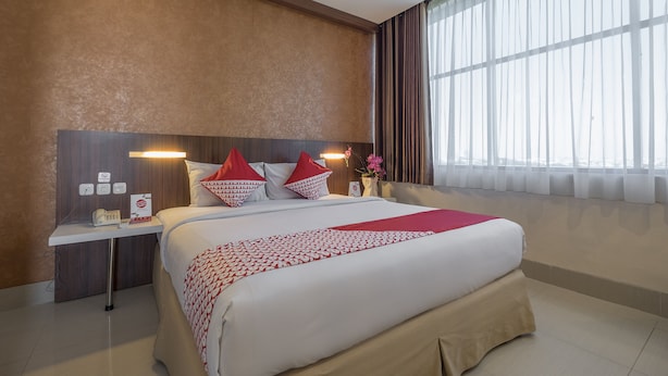 A hotel room with a large bed, a bedside table with a lamp, a clock, and a vase with flowers. - All Nite and Day Makassar (Makassar)