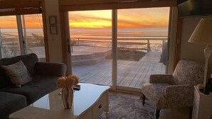 A living room with a large sliding glass door leading to a wooden deck overlooking the beach at sunset. - Oceanfront w/Private Stairs to Beach and Spectacular Sunsets! (Eastham)