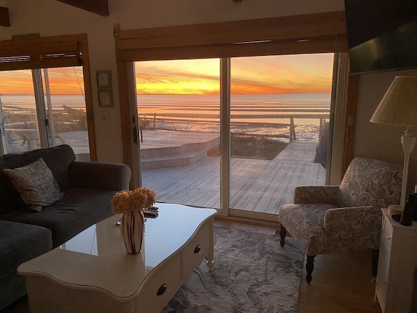 Un salon doté d’une grande porte-fenêtre coulissante qui donne sur une terrasse en bois offrant une vue sur la plage au coucher du soleil.