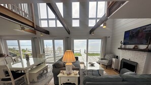 A spacious living room with a large glass door leading to a deck, a fireplace, and a television mounted on the wall. - Oceanfront w/Private Stairs to Beach and Spectacular Sunsets! (Eastham)