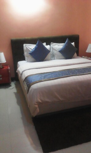 A neatly made bed with white bedding and blue pillows, a red nightstand with a lamp, and a black headboard. - Home Inn Hotels and Suites (Lagos)