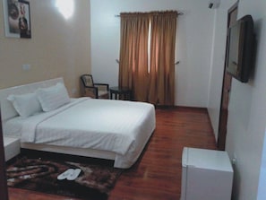 A small, neatly arranged bedroom with a bed, a chair, a small table, and a TV mounted on the wall. - Newcastle Hotel Lekki (Lagos)