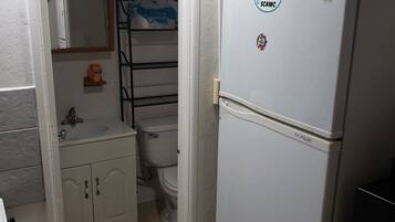 A compact kitchen with a refrigerator, a sink, and a toilet visible through an open door.