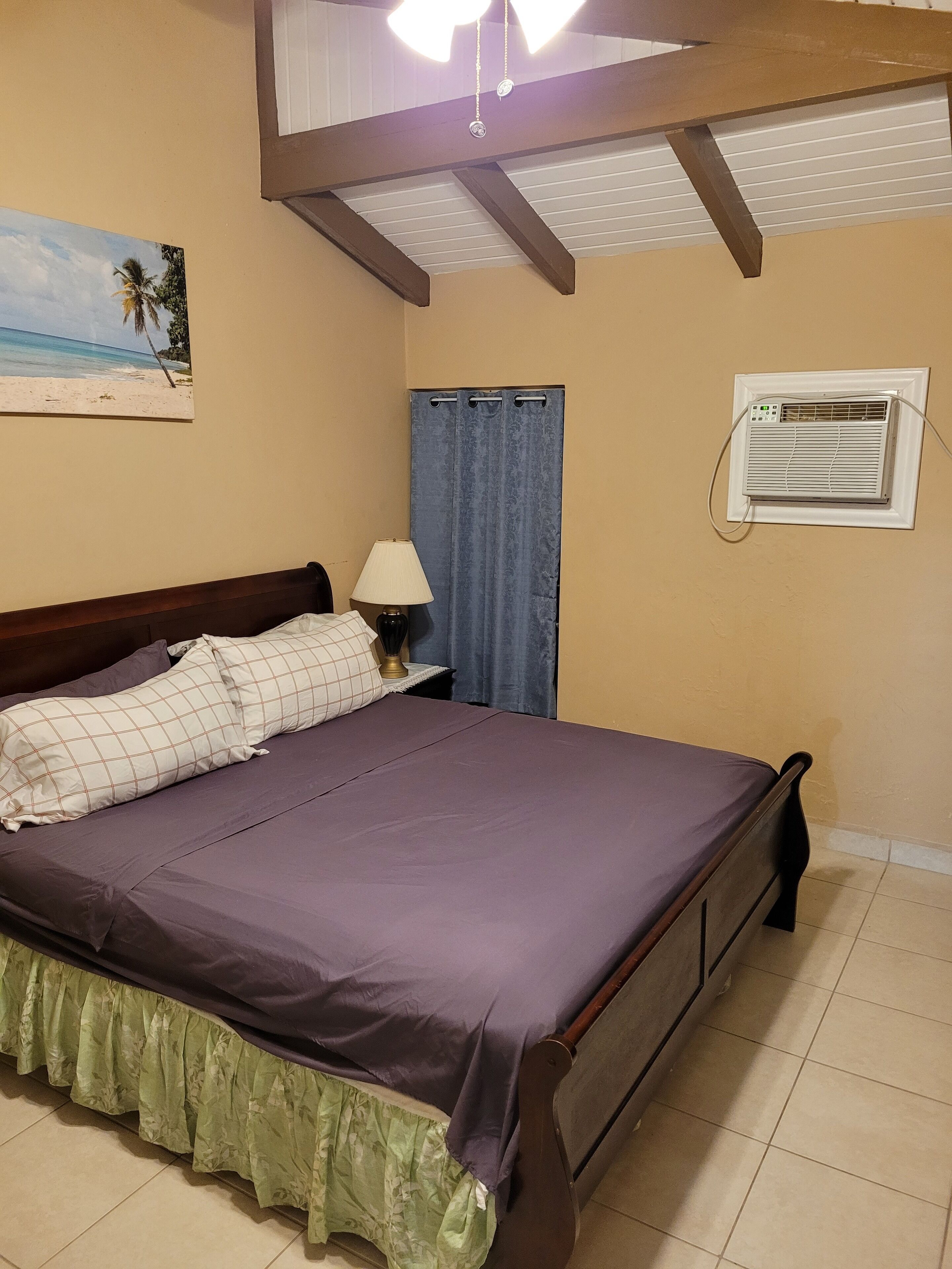 A bedroom with a bed, a nightstand with a lamp, a window with curtains, and an air conditioning unit.