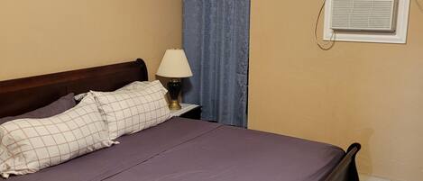 A bedroom with a bed, a nightstand with a lamp, a window with curtains, and an air conditioning unit.