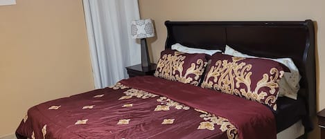 A neatly made bed with a maroon bedspread and matching pillows, a ceiling fan, a wall-mounted lamp, and two floral paintings.