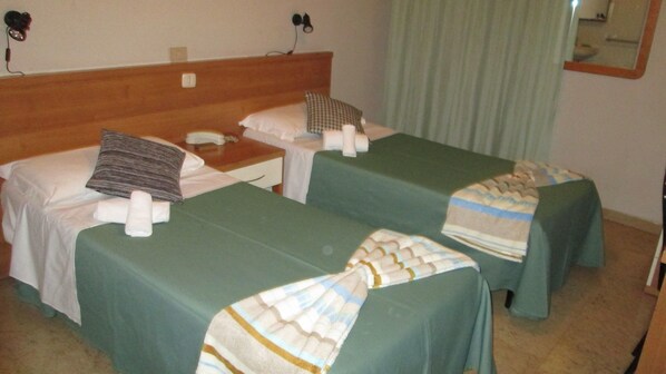 A hotel room with two single beds, each with a pillow and a blanket. There is a wooden headboard, a nightstand with a lamp, and a mirror on the wall.