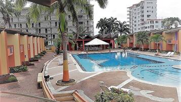 A swimming pool with lounge chairs, a covered seating area, and a building with balconies in the background.
