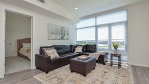 A modern living room with a sectional sofa, a coffee table, and a rug. There is a window with blinds, a ceiling fan, and a painting on the wall. - Contemporary Urban Living in Gated Complex |2Q (Katy)