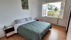 A bedroom with a bed, a nightstand, a window with a view of greenery, and a radiator. - MAISON DE MARITXU - Between beach and forest - Landes - Pays Basque (Tarnos)