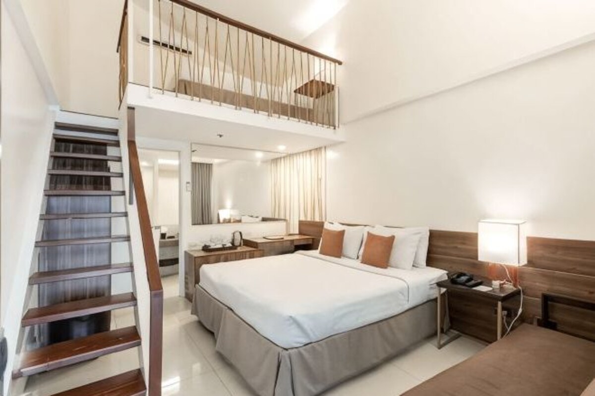 A hotel room with a bed, bedside tables, a nightstand, a lamp, a wooden staircase, and a balcony.