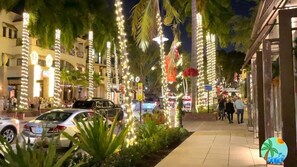 A street lined with palm trees and illuminated by string lights, with parked cars and pedestrians. - Enjoy a new construction home with heated salt pool close to everything Naples (Naples)