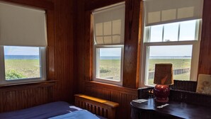 Ett rum med väggar beklädda med träpaneler, en säng med ett blått täcke, ett litet bord med en lampa och stora fönster med utsikt över stranden.