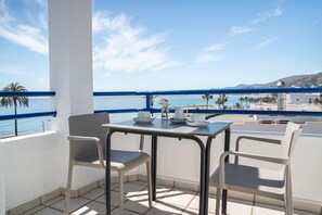 A balcony with a table and chairs, overlooking a coastal view with palm trees and a clear sky.