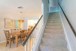Una escalera con barandilla, un área de comedor con sillas de madera y una mesa, y una cocina al fondo.