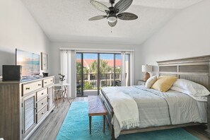 A bedroom with a bed, a ceiling fan, a TV, a desk, and a view of the outdoors.