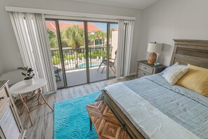 Habitación de hotel con cama, escritorio, silla, mesita con planta en maceta, alfombra azul estampada y vista a una piscina y palmeras al exterior.