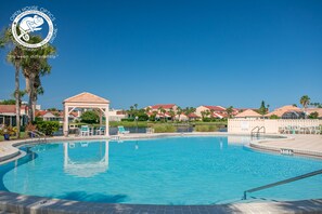 A swimming pool with lounge chairs and a gazebo, surrounded by palm trees and houses with red roofs.