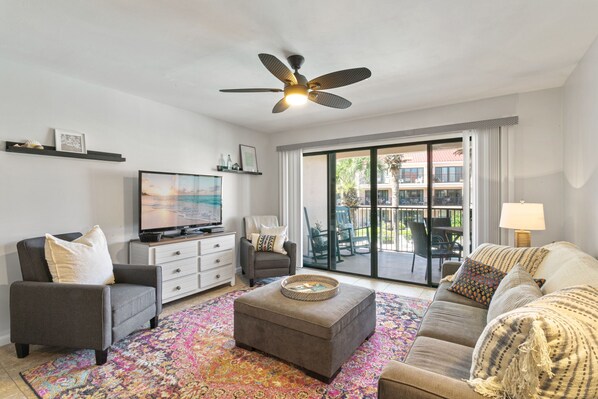 A living room with a sofa, armchair, ottoman, TV, ceiling fan, and a view through a glass door to an outdoor patio.