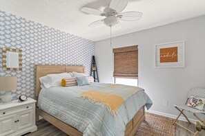 Un dormitorio con cama, mesita de noche, ventilador de techo, ventana con persianas y un cuadro en la pared.