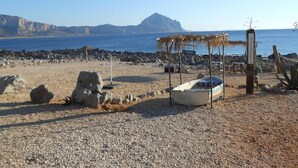 A beach with a small boat, a thatched shelter, and rocky terrain. - Happy Camp in El Bahira Camping Village (San Vito Lo Capo)
