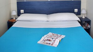 A neatly made bed with a blue bedspread and white pillows, a bedside table with a lamp, a shelf above the bed with books and a plant, and a magazine on the bed. - Happy Camp in El Bahira Camping Village (San Vito Lo Capo)