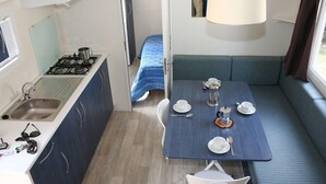 A compact kitchenette with a gas stove, sink, and a small window. A dining area with a table set for two, a bench, and a hanging light fixture is visible. - Happy Camp in El Bahira Camping Village (San Vito Lo Capo)