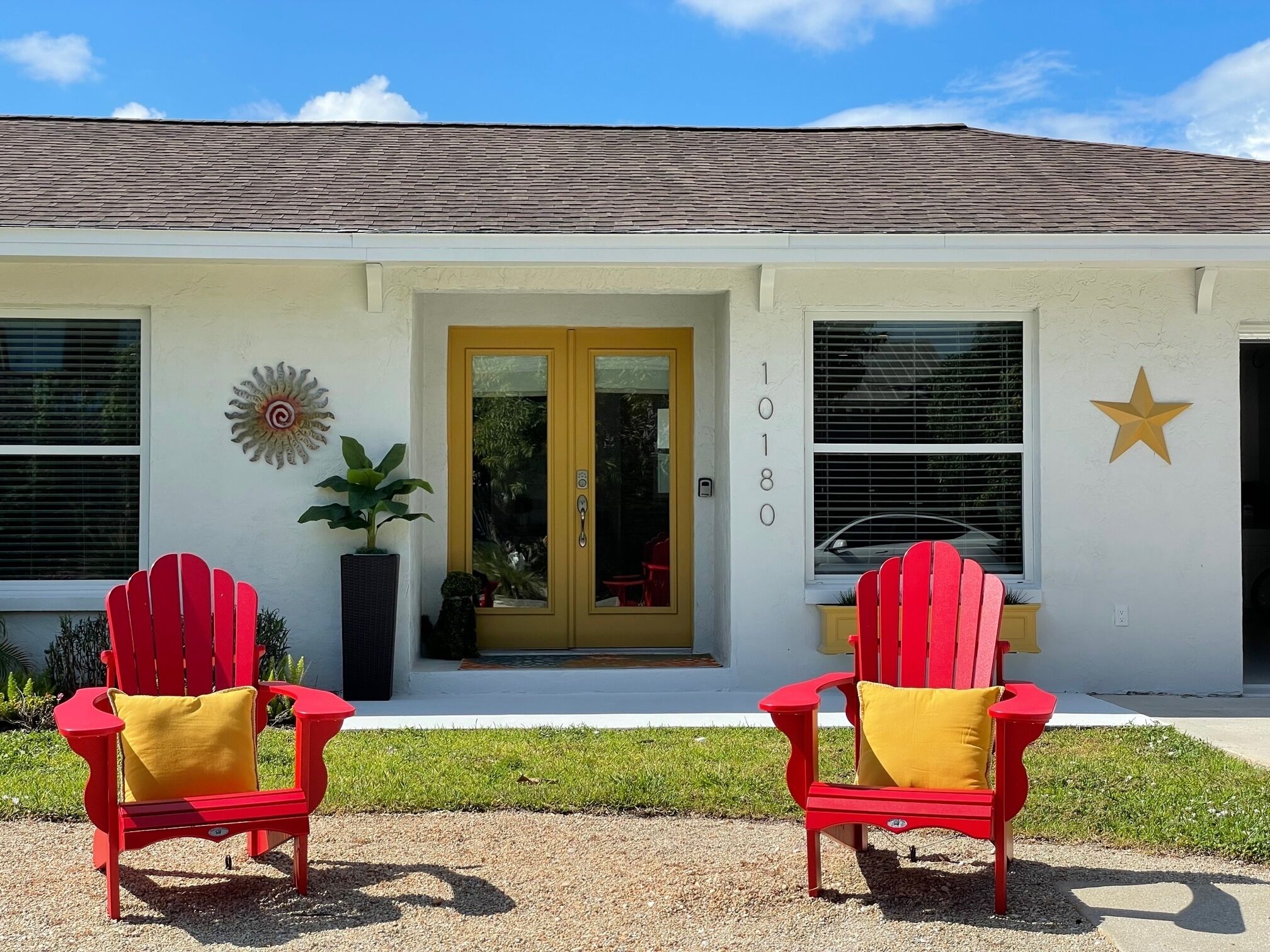 A white building with a yellow door, two red Adirondack chairs with yellow cushions, a star decoration, and the number 1018 on the wall.