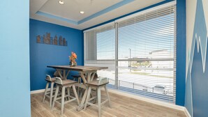 Ein Zimmer mit blauer Wand, einem Holztisch mit vier Stühlen, einer Vase mit Blumen und einem großen Fenster mit Jalousien.