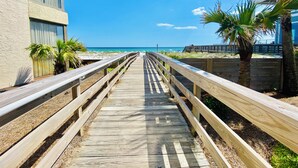Une promenade en bois menant à une plage, bordée d’immeubles et de palmiers.