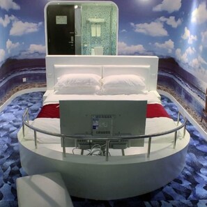 A circular bed with a white mattress and pillows, a small bedside table, and a wall-mounted TV. - Grand Nest Hotel (Zhuhai)