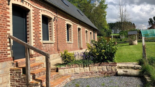 Un bâtiment en briques avec un toit en pente, un petit jardin avec du gravier et des plantes, et un escalier en bois menant à l’entrée.