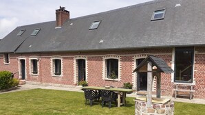 Une maison en briques avec un toit en pente, un puits en pierre et une terrasse avec une table et des chaises.