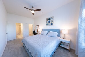 A bedroom with a bed, a nightstand, a desk, a chair, a ceiling fan, and a painting on the wall. - Spacious Private Condo Unit in Lifestyle Center (Katy)