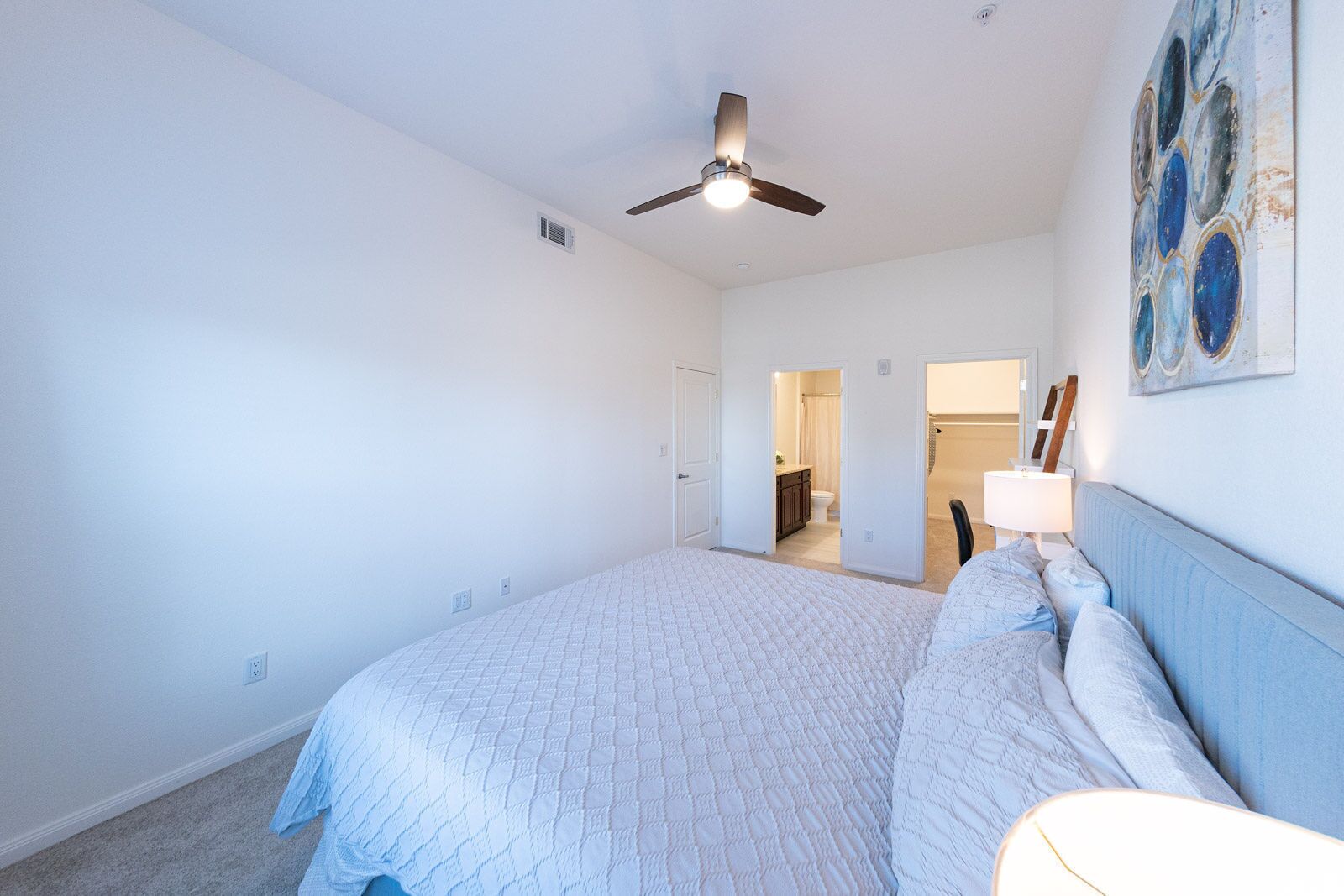 Ein Schlafzimmer mit einem Bett, einem Deckenventilator, einem Gemälde und einem durch eine geöffnete Tür einsehbaren Badezimmer.