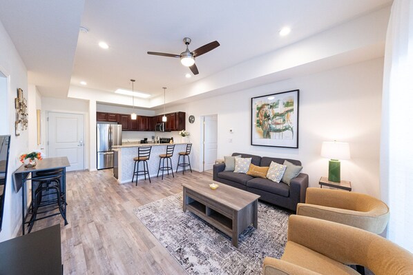 A modern living room with a sofa, coffee table, and bar stools. There is a kitchen area with a refrigerator, microwave, and island. - Spacious Private Condo Unit in Lifestyle Center (Katy)