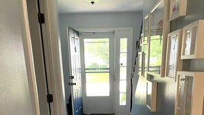 Een gang met houten vloer, witte muren en een deur naar een lichte kamer met ramen.