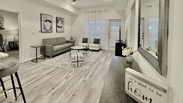 A modern living room with a sofa, chairs, a coffee table, and a ceiling fan. There is a sign on the wall that reads "It's good to be home." - 1K| Spacious Home in Gated Complex| Walk to your Daily Needs (Katy)