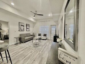 Una sala de estar moderna con un sofá, dos sillones, una mesa de centro y un ventilador de techo. Hay un espejo en la pared y un cartel con texto.