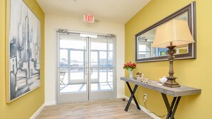 A hallway with a yellow wall, a large abstract painting, a door with glass panels, and a table with a lamp and vase. - 1K| Spacious Home in Gated Complex| Walk to your Daily Needs (Katy)