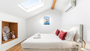 A modern bedroom with a skylight, a bed with white bedding and red pillows, a bedside table with a lamp, and a wall-mounted air conditioner. - GuestReady - Freixo Island - Douro Views (Porto)