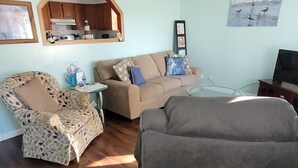 Una sala de estar con un sofá, un sillón y un reposapiés. Hay una zona de cocina con armarios de madera y un microondas. Un cuadro enmarcado cuelga de la pared.