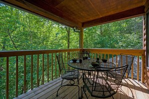 Une terrasse extérieure couverte, dotée d’une table ronde et de chaises, entourée d’une balustrade en bois, et offrant une vue sur une forêt dense.